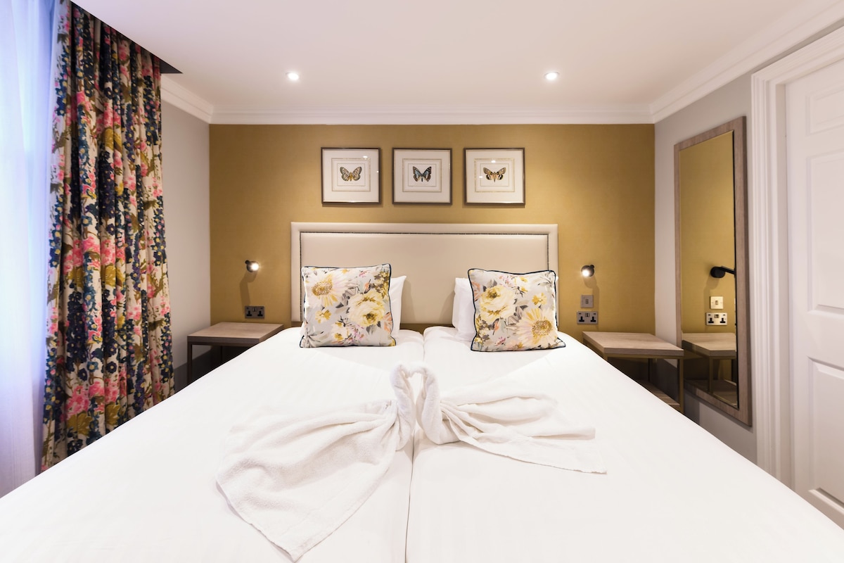 A hotel room with a large bed, floral-patterned pillows, a headboard, bedside tables with lamps, and a mirror on the wall.