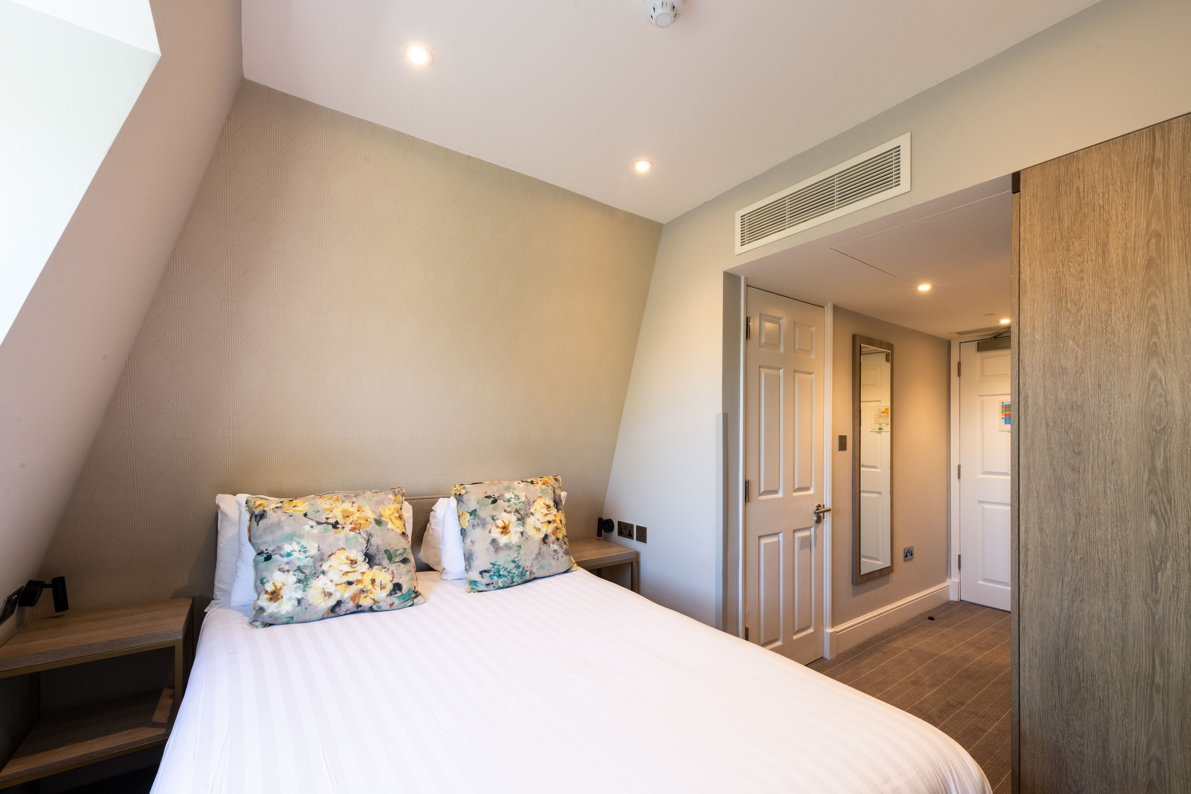 A bedroom with a bed, two floral pillows, a nightstand, a wooden wardrobe, and a door leading to another room.