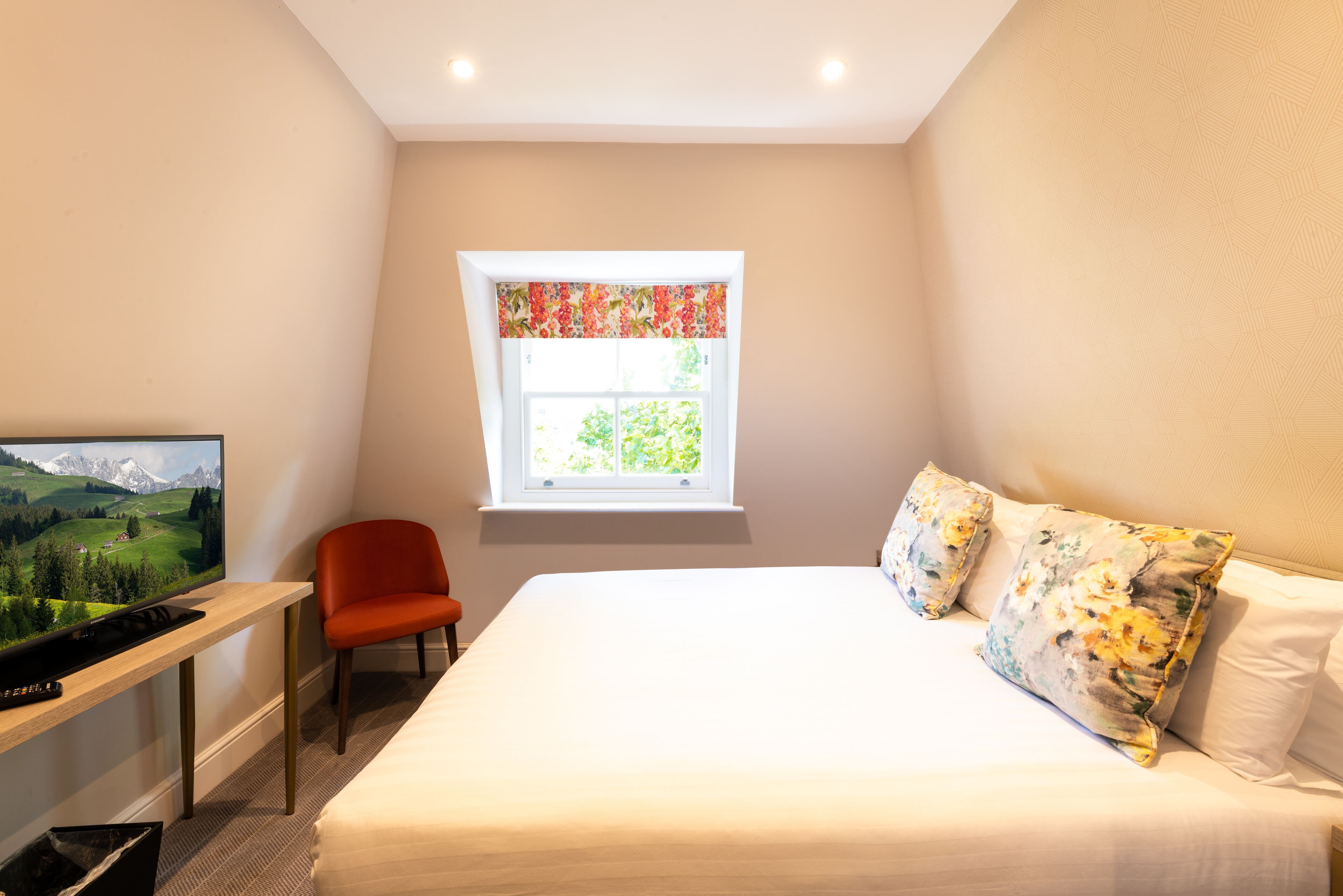 Un dormitorio con cama, una televisión sobre un escritorio, una silla y una ventana con vistas a zonas verdes.