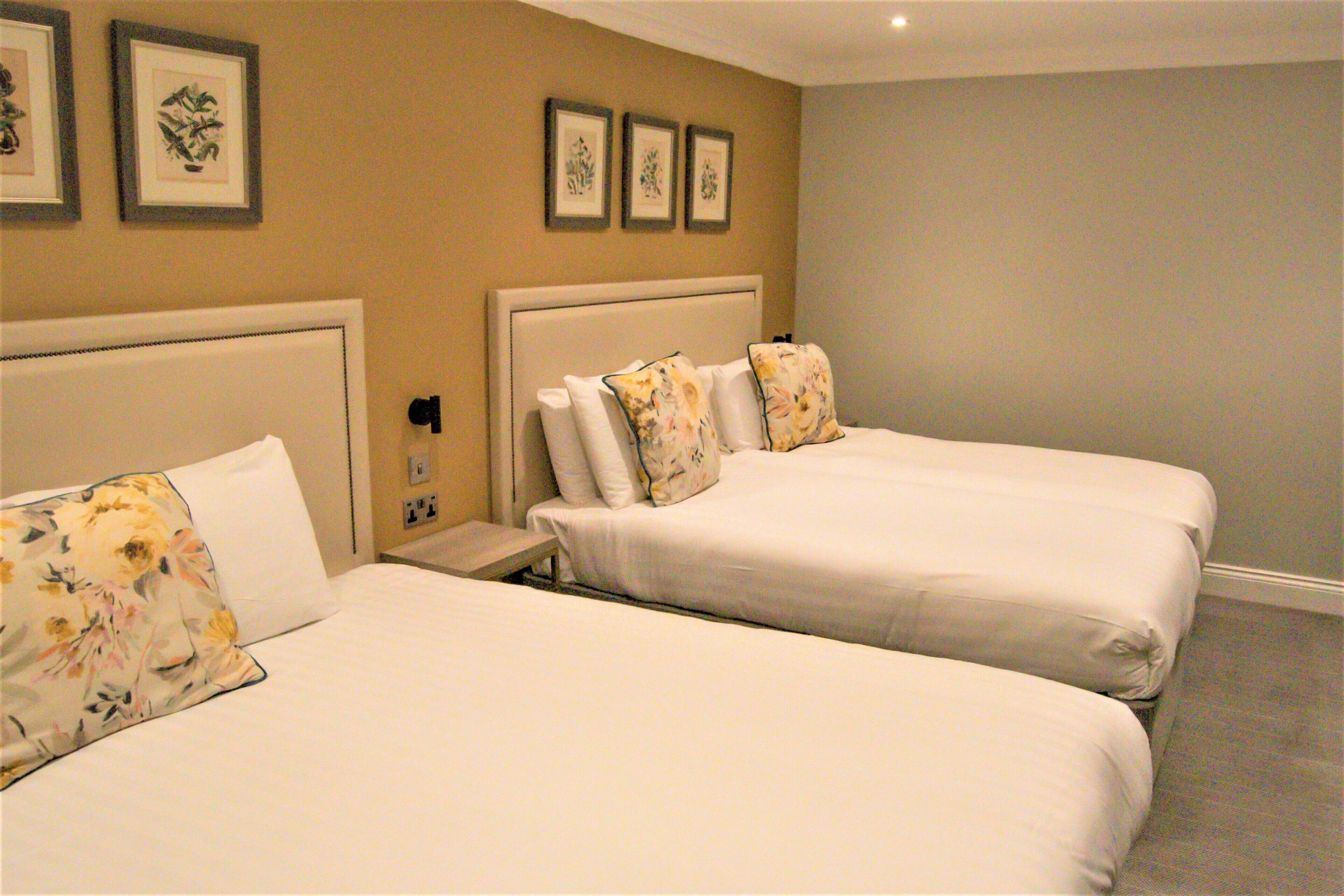 Una habitación de hotel con dos camas, cada una con almohadas de estampado floral, una mesita de noche y una obra de arte enmarcada en la pared.