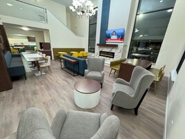 Un vestíbulo de hotel moderno con zona de comedor, asientos, chimenea y una televisión mostrando noticias.