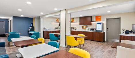 A modern hotel room with a kitchenette, a dining area with tables and chairs, and a separate living area with a sofa.