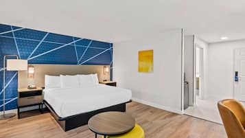 Habitación de hotel con cama, mesitas de noche, lámpara, una pequeña mesa redonda, un otoman amarillo y un diseño geométrico azul en la pared.