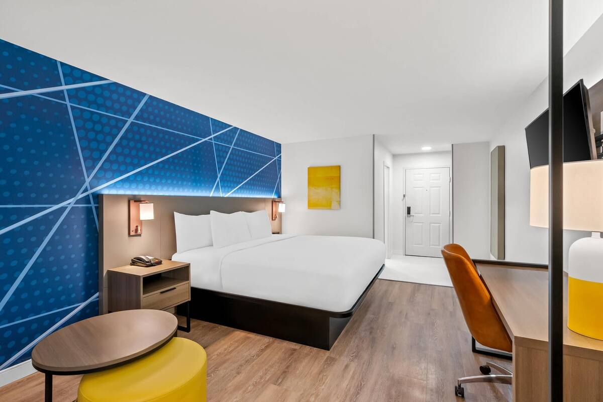 A modern hotel room with a large bed, a desk, a chair, and a small table.