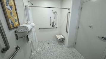 A bathroom with white tiled walls, a shower with grab bars, a toilet with a safety bar, and a towel rack with folded towels.