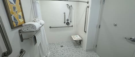 Ein Badezimmer mit weißen Fliesen, einer Dusche mit Haltegriffen, einer Toilette mit Sicherheitsbügel und einem Handtuchhalter mit gefalteten Handtüchern.