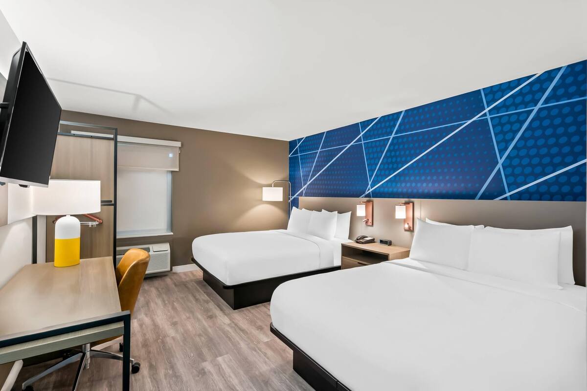 A hotel room with two beds, a desk, a chair, a TV, and a wall with a geometric pattern.