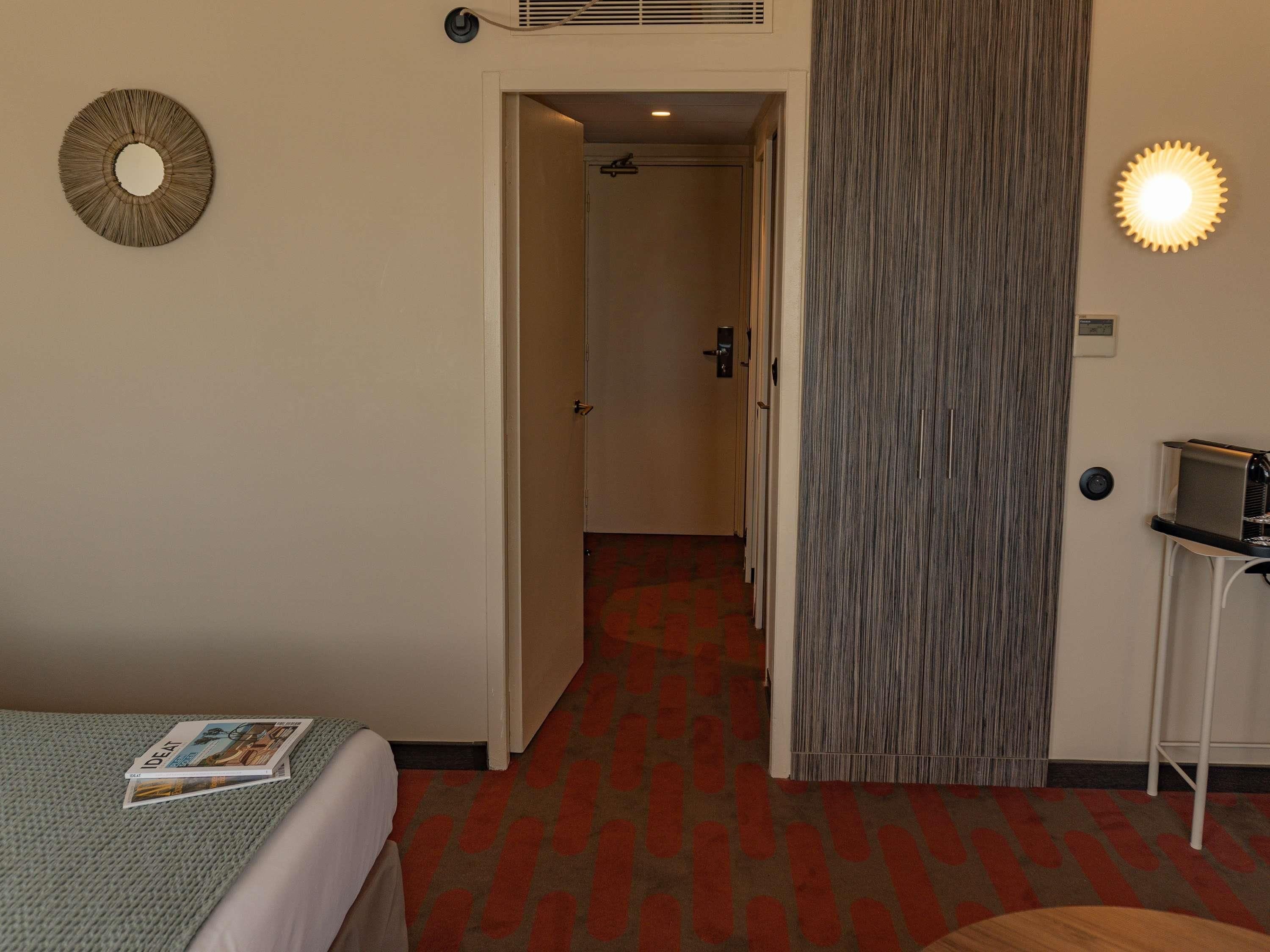 A hotel room with a bed, a door leading to a hallway, a wall-mounted light, and a small table with a coffee machine.