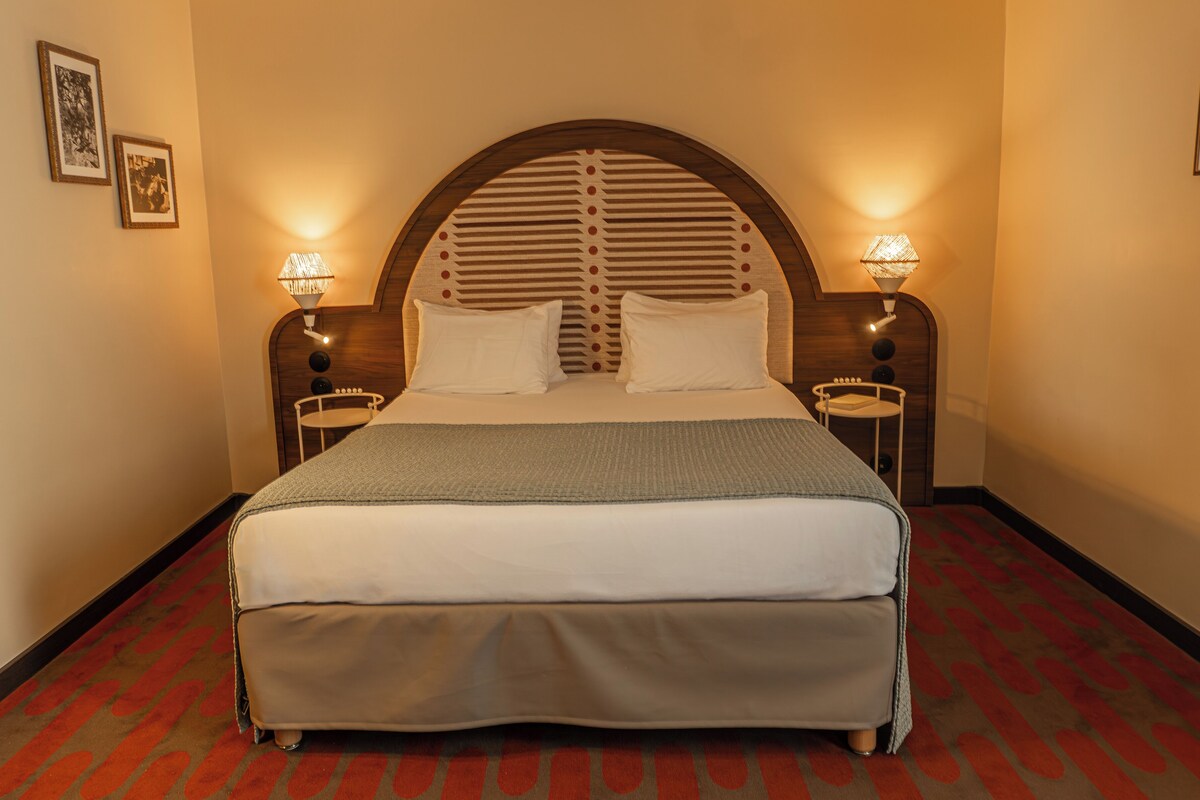 Una cama bien hecha con cabecera de madera, dos mesitas de noche con lámparas y una alfombra estampada.