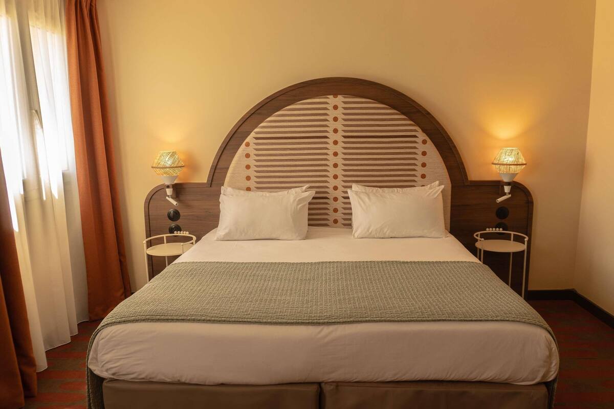 A hotel room with a large bed, two bedside lamps, a headboard with a geometric pattern, and a window with curtains.