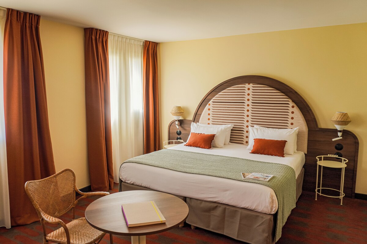 Habitación de hotel con una cama grande, dos mesitas de noche, una mesa redonda, una silla de mimbre y vistas al exterior a través de cortinas.