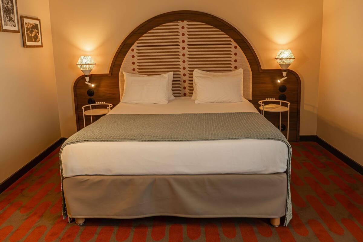 A hotel room with a large bed, two bedside tables with lamps, a headboard with a unique design, and a patterned carpet.