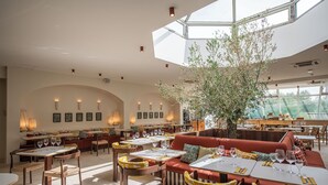 A spacious restaurant with high ceilings, multiple dining tables, and a central seating area with a tree. - Mercure Arles Centre Arenes (Arles)