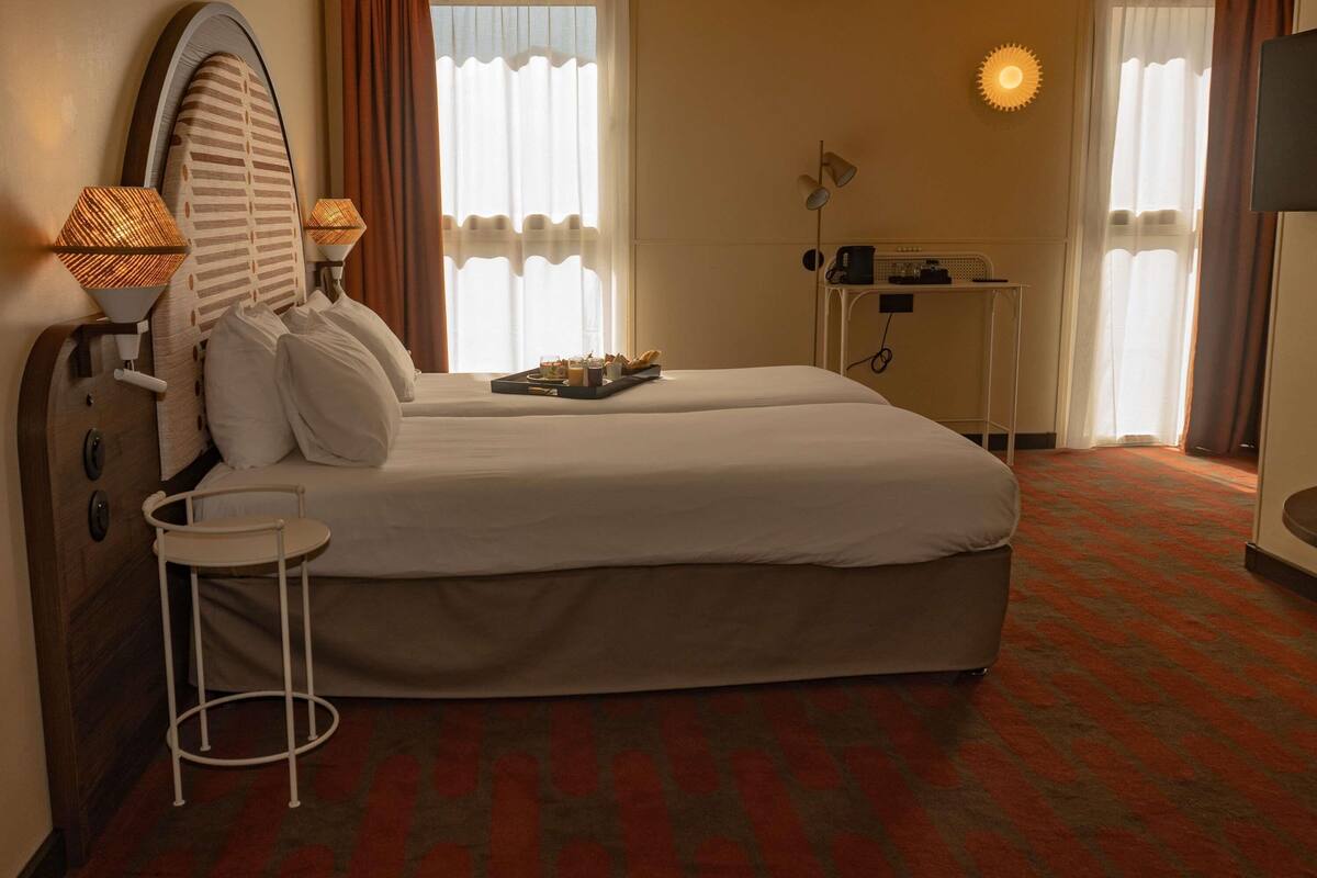 A hotel room with two beds, a bedside table, a lamp, a desk with a chair, and a TV.