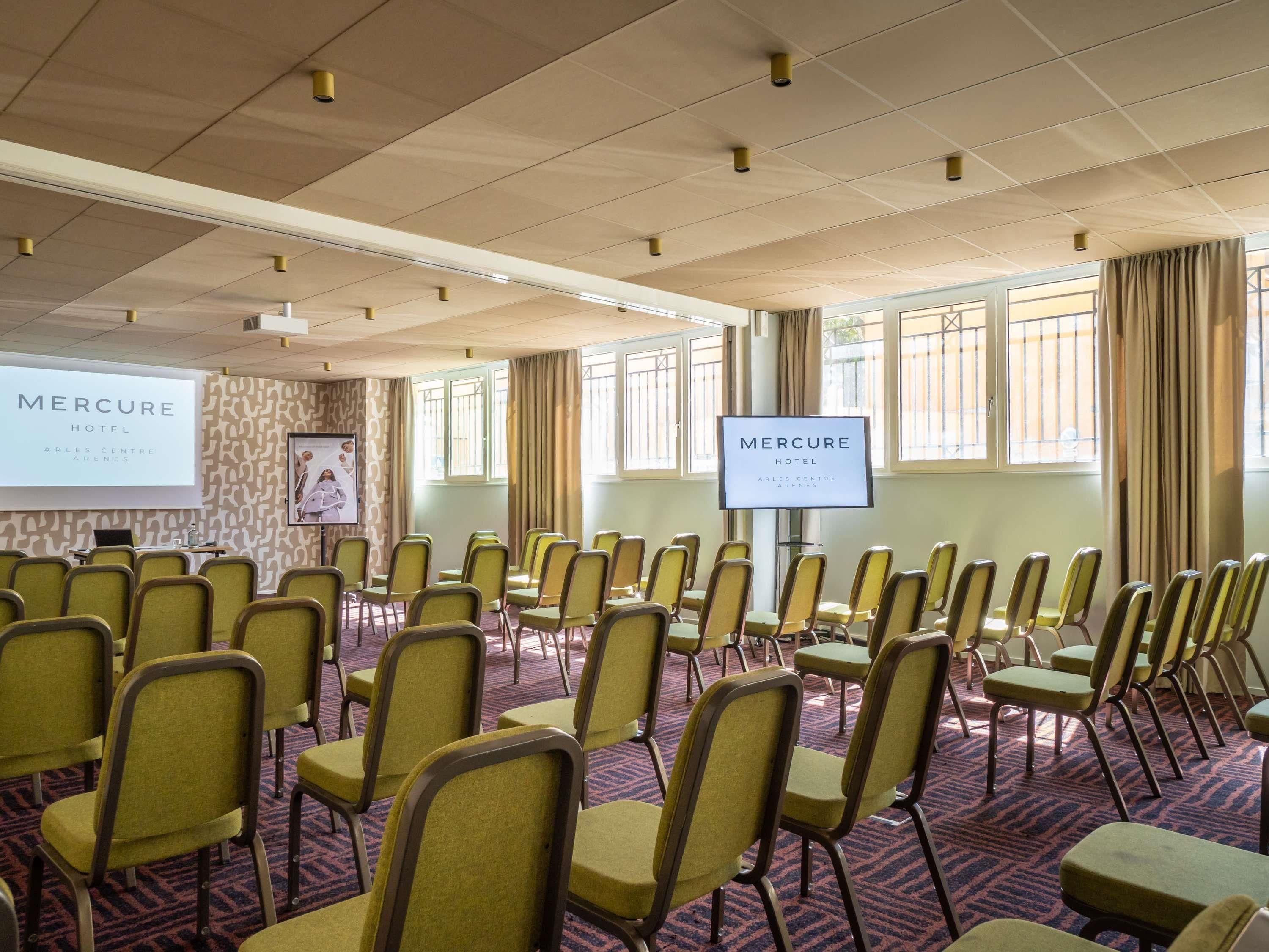 A conference room with rows of chairs, a projection screen, and a television screen displaying the hotel name.