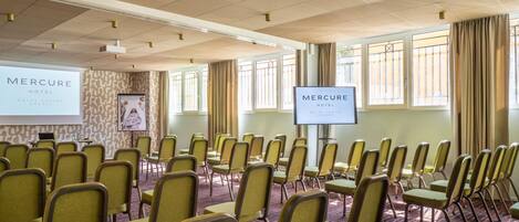 A conference room with rows of chairs, a projection screen, and a television screen displaying the hotel name.