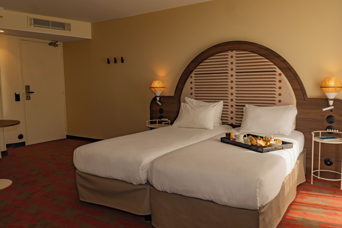 Una habitación de hotel con una cama grande, dos mesitas de noche con lámparas, una mesita pequeña y una puerta.
