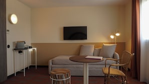 Una habitación de hotel con un sofá, dos sillones, una mesita, un televisor montado en la pared, un escritorio con una impresora y una lámpara.