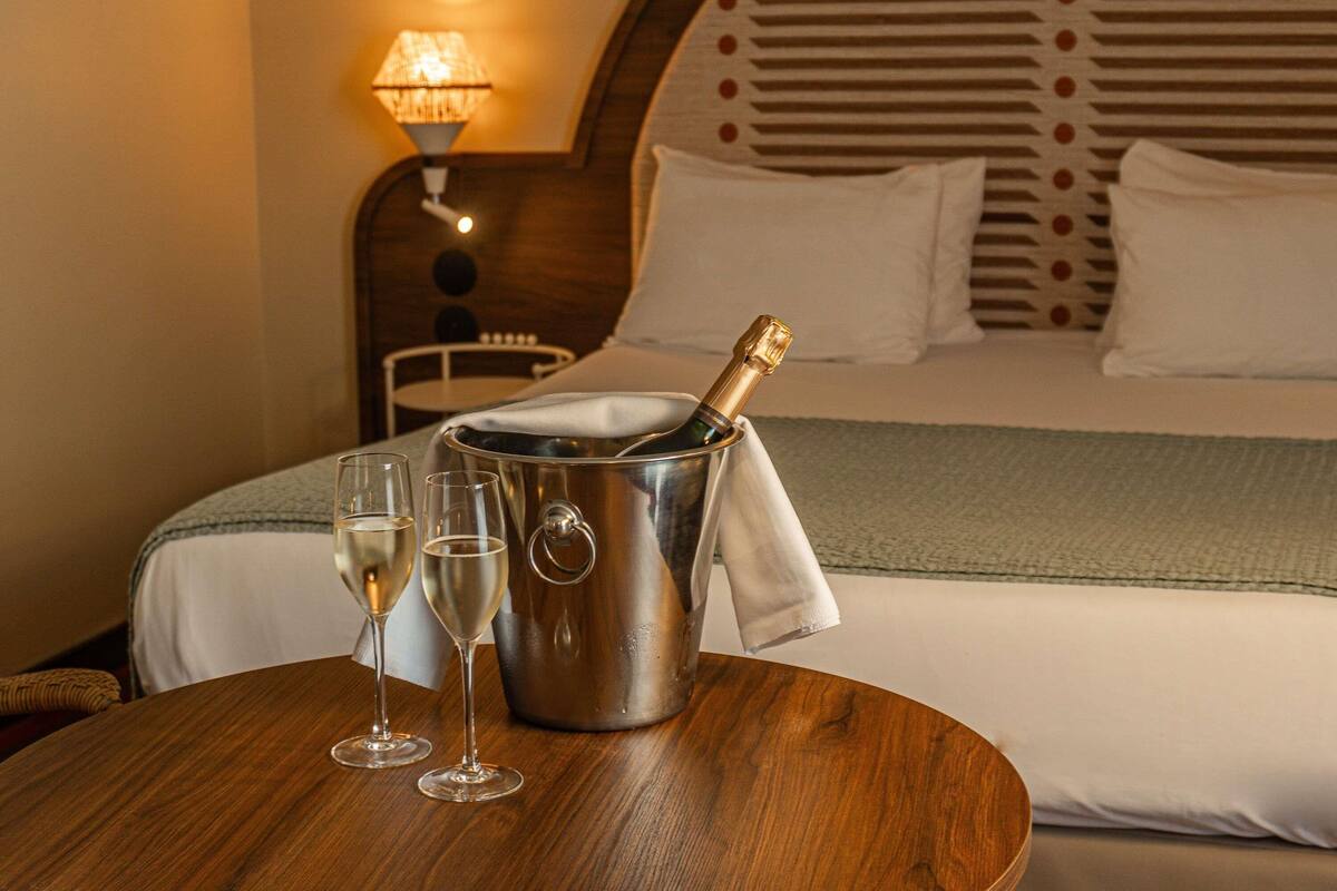Quarto de hotel com cama, mesa de cabeceira com uma garrafa de champanhe e copos, luminária e cabeceira estampada.