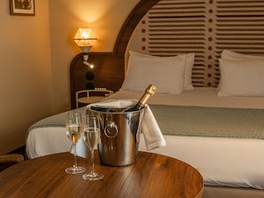 A hotel room with a bed, a bedside table with a champagne bottle and glasses, a lamp, and a patterned headboard.