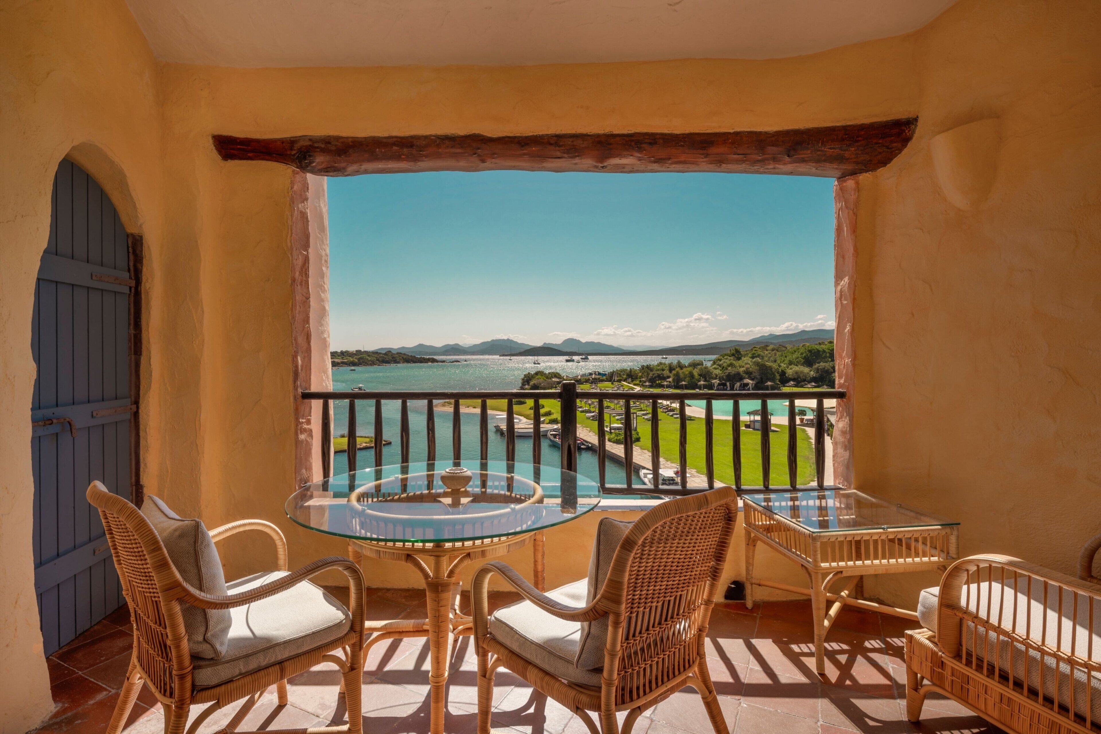 Ein Balkon mit Korbmöbeln, einem Glastisch und Blick auf ein Gewässer sowie viel Grün.