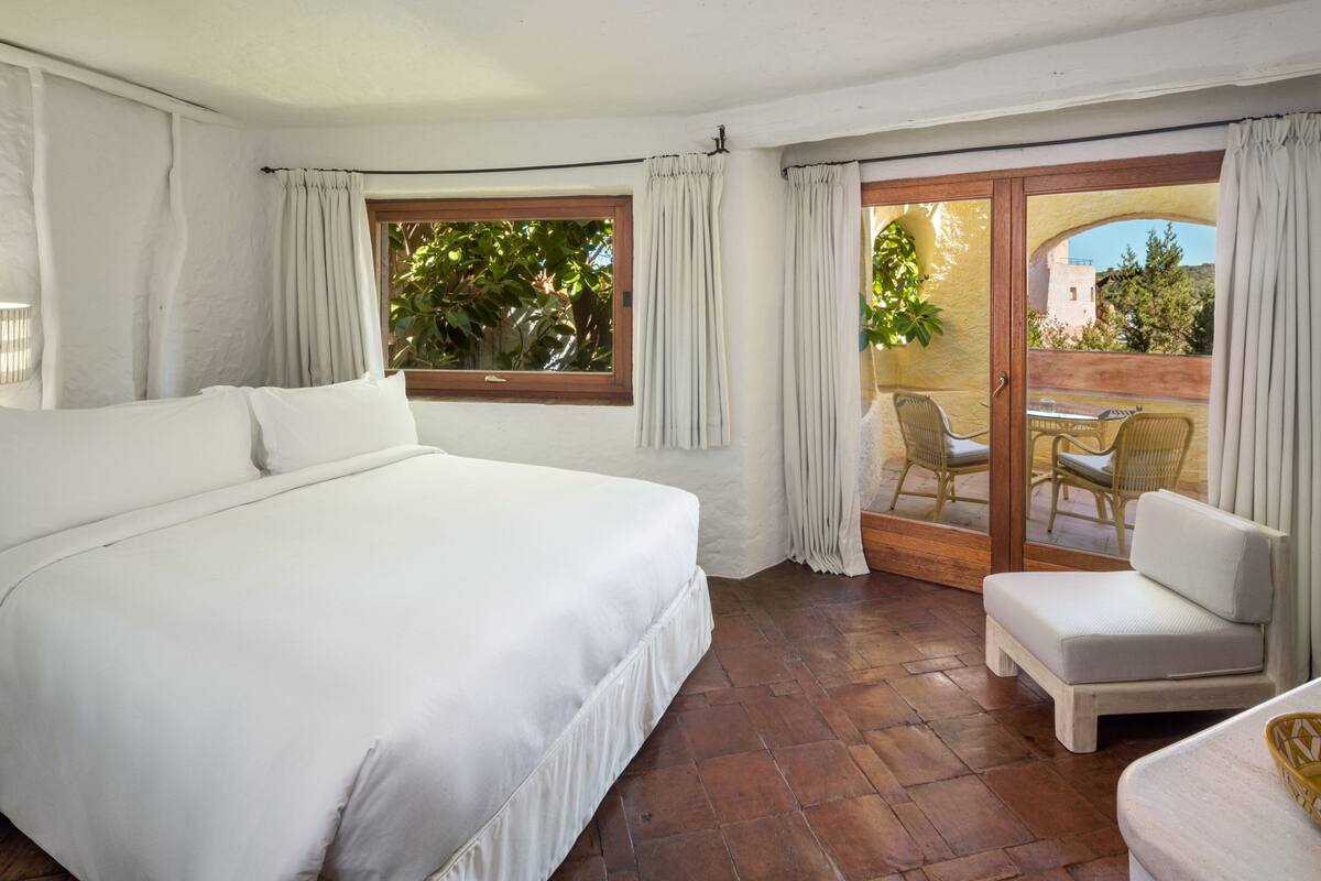 Une chambre avec un grand lit, un balcon doté de fauteuils en osier et une vue sur la verdure et les bâtiments.