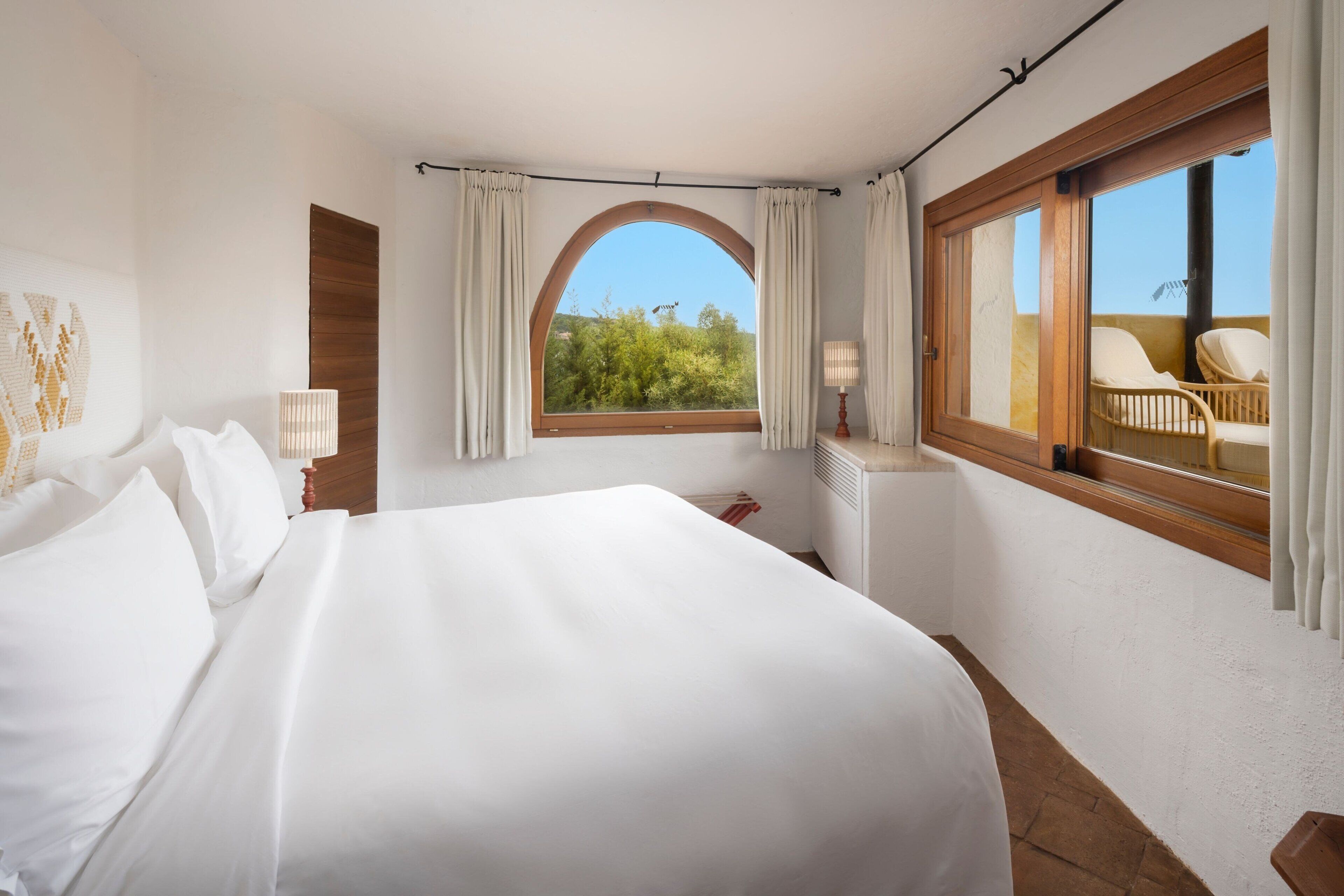 A bedroom with a large bed, a window with curtains, a bedside table with a lamp, and a view of the outdoors.