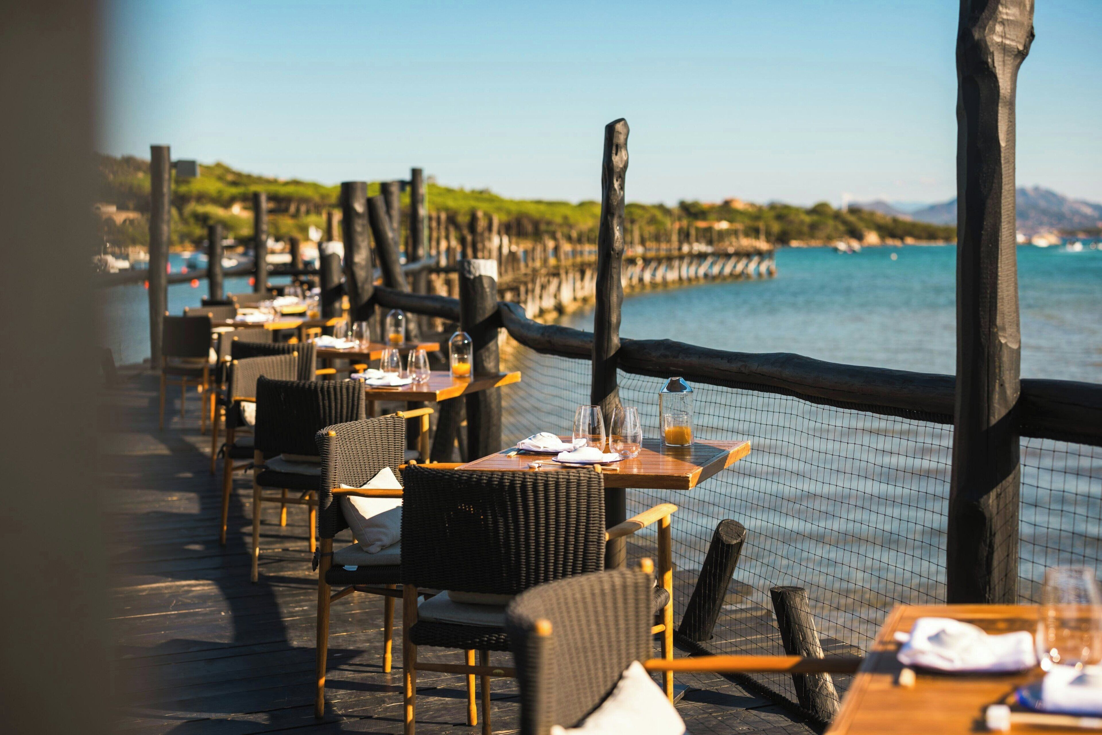 Un área de comedor al aire libre con sillas y mesas de mimbre, con vistas a un cuerpo de agua y una costa distante.