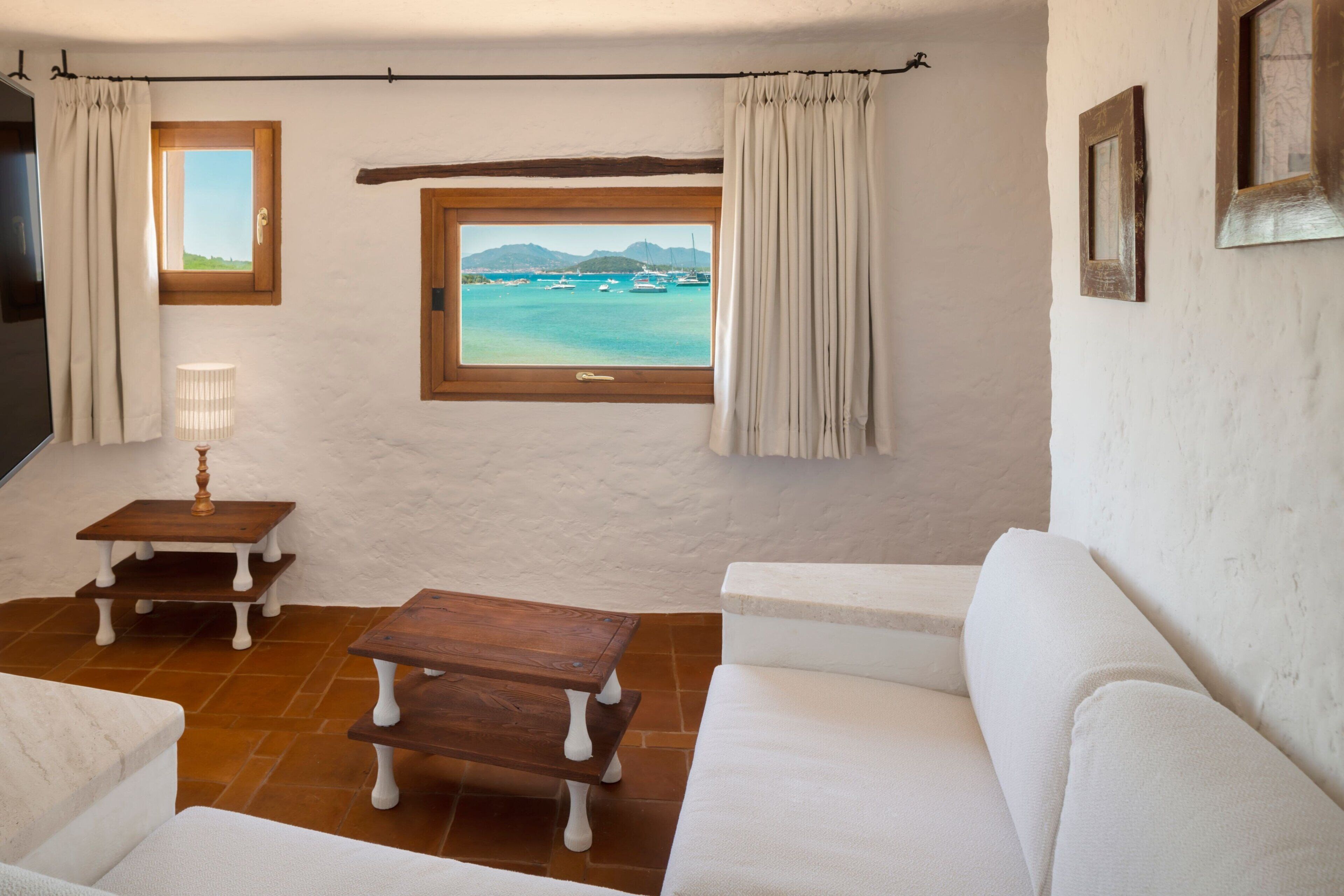 A room with a white couch, a wooden coffee table, a small side table, a window with a view of the sea, and a door.