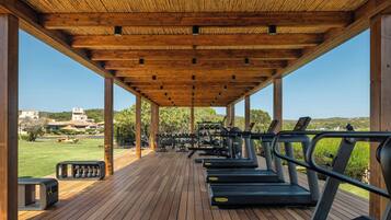 Zona de gimnasio techada al aire libre con equipo de ejercicio, incluyendo cintas de correr y máquinas de pesas, bajo un techo de madera.