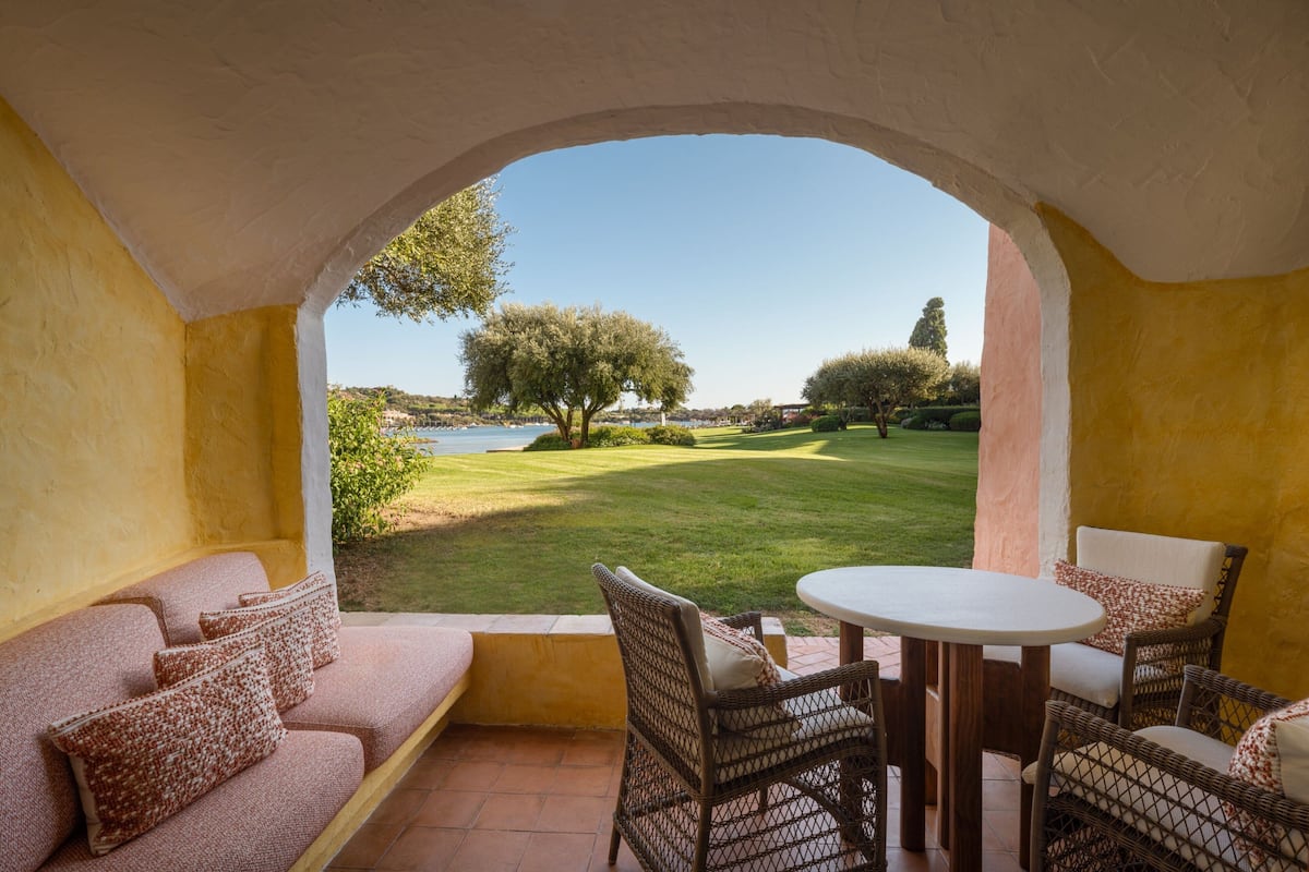 Une terrasse avec une table ronde et des chaises en osier, un canapé avec des coussins à motifs et une vue sur un espace verdoyant, avec des arbres et une étendue d’eau.