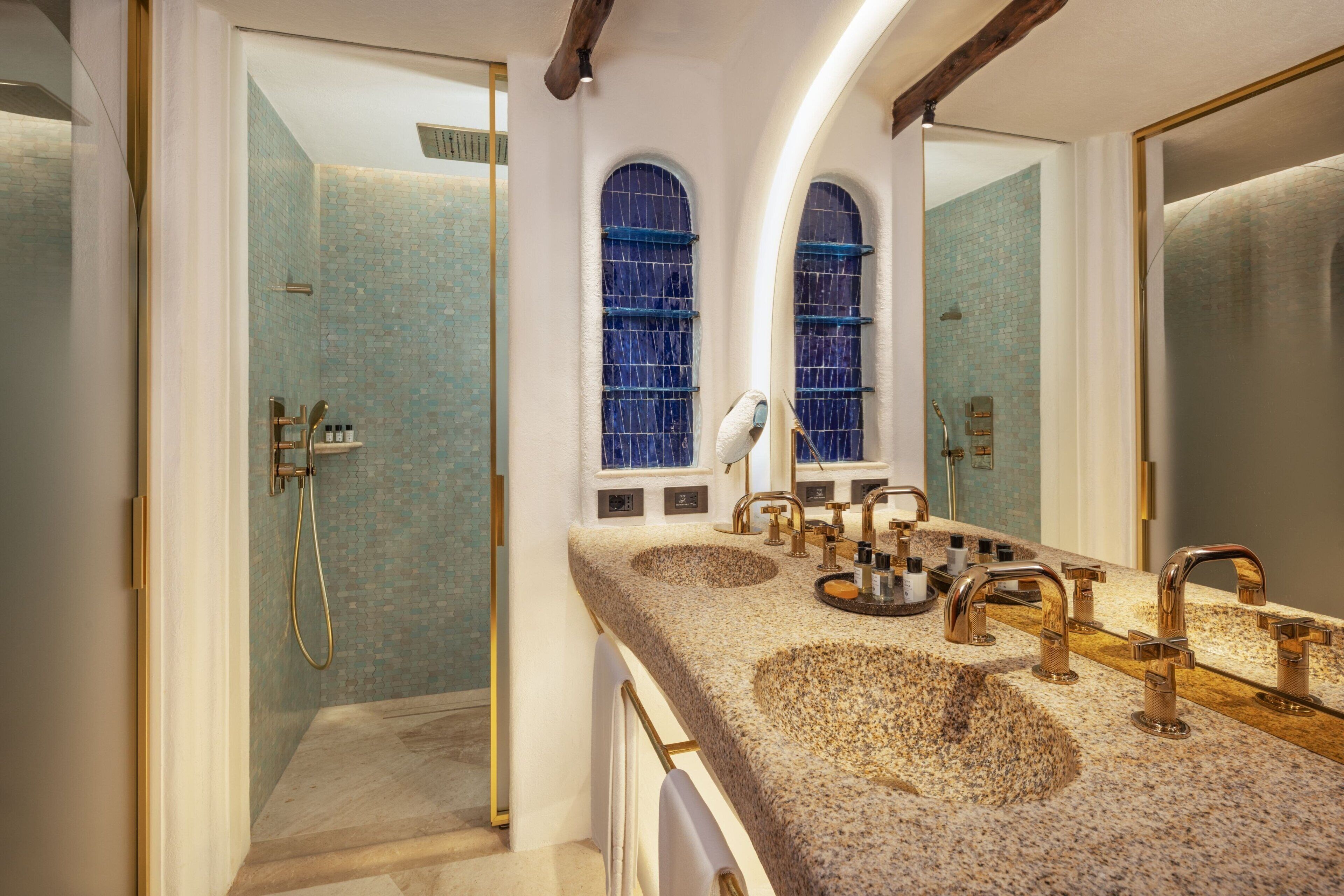 A bathroom with a large mirror, a shower with a glass door, and a granite countertop with multiple sinks and faucets.