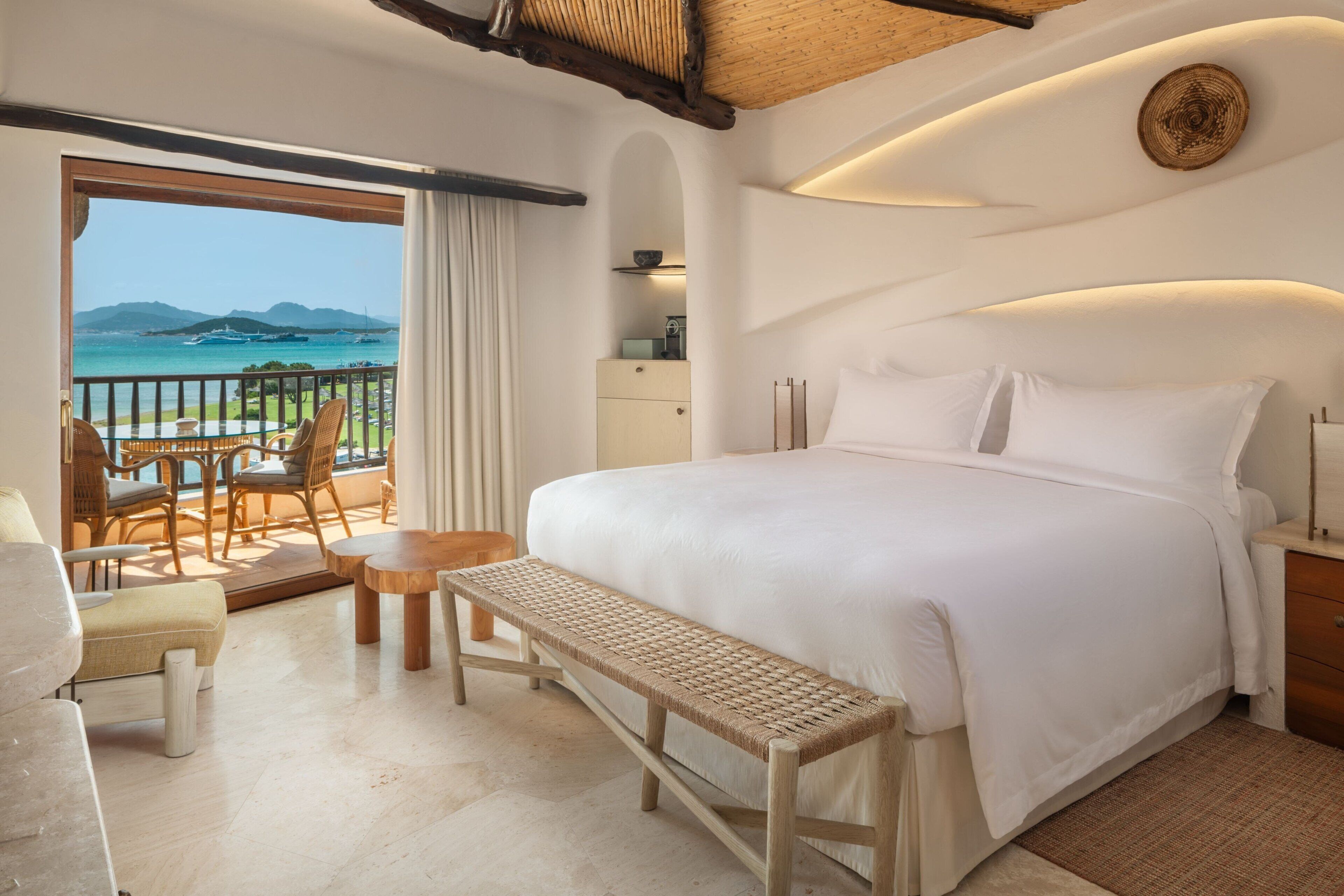 Ein Hotelzimmer mit einem großen Bett, einem kleinen Tisch, einer Bank und Blick aufs Meer.
