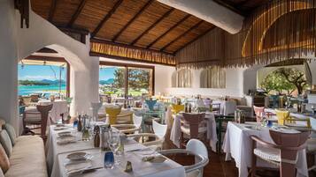 Un restaurant doté d’une terrasse offrant une vue sur la côte. Les tables sont dressées avec des nappes blanches et accueillent différents plats.
