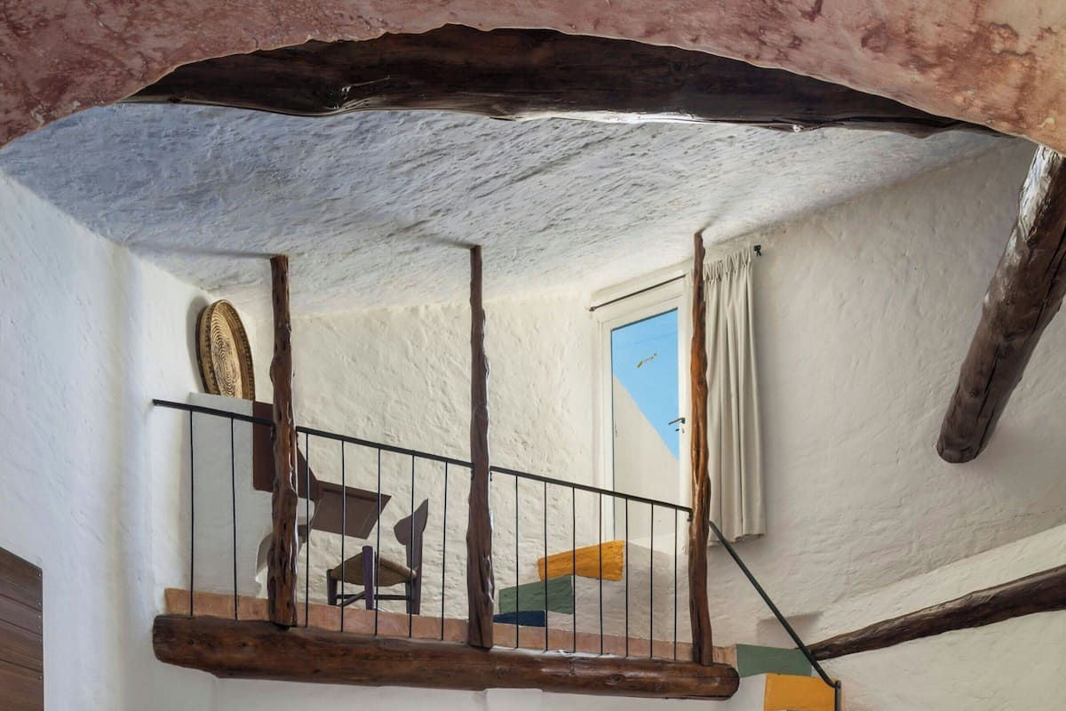 Une chambre à coucher avec un grand lit, une chaise en bois, une table de chevet, une lampe, un escalier aux marches colorées et une vue sur le ciel par une fenêtre.