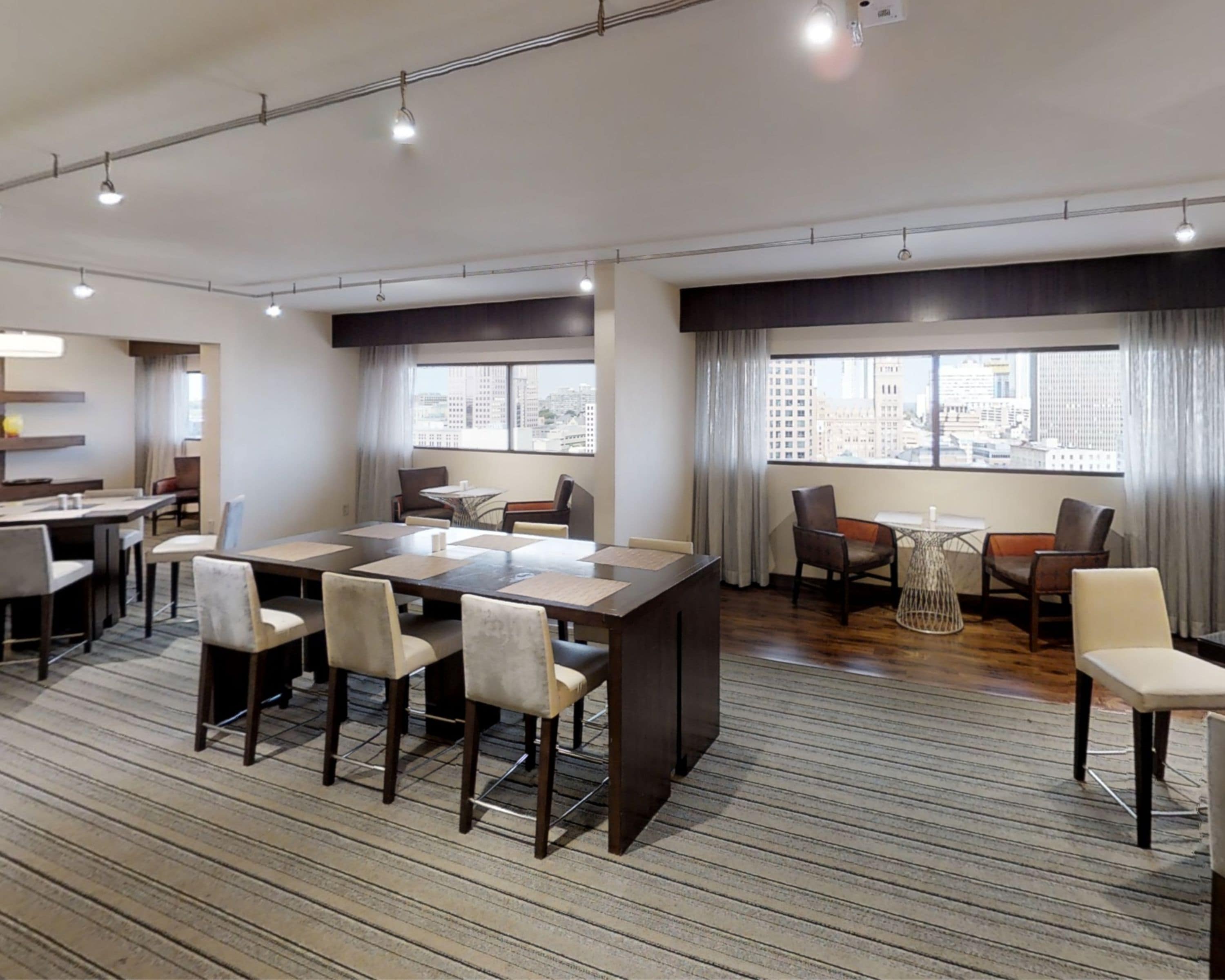 A modern hotel room with a large dining table, comfortable seating, a small kitchen area, and large windows offering a city view.