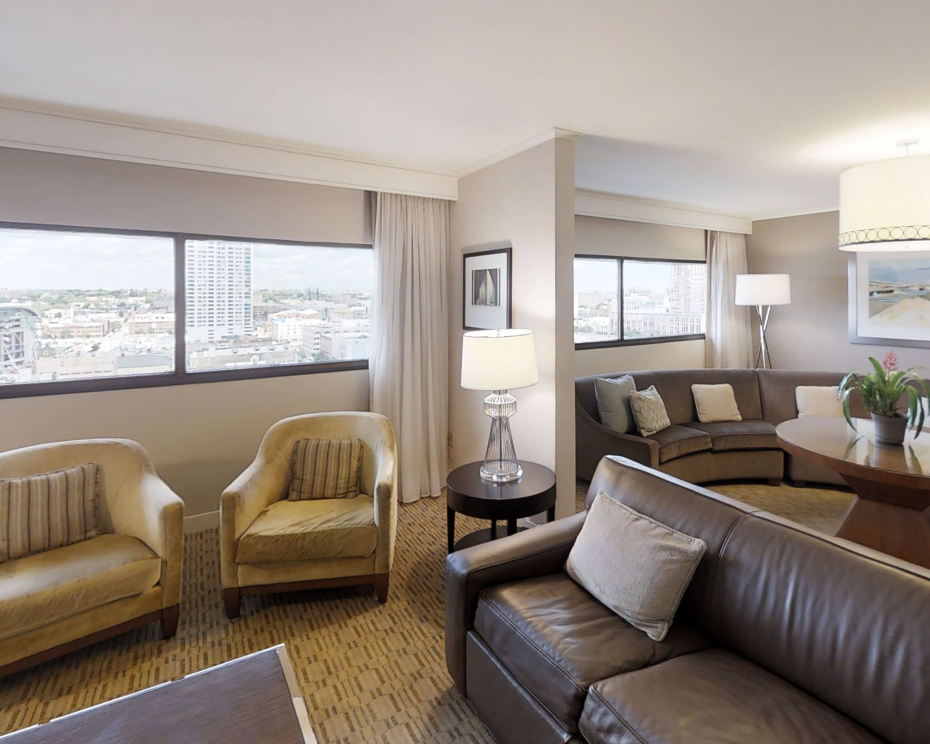 Quarto de hotel com vista para a cidade, um sofá, duas poltronas, uma mesinha de centro com uma luminária e uma janela grande.