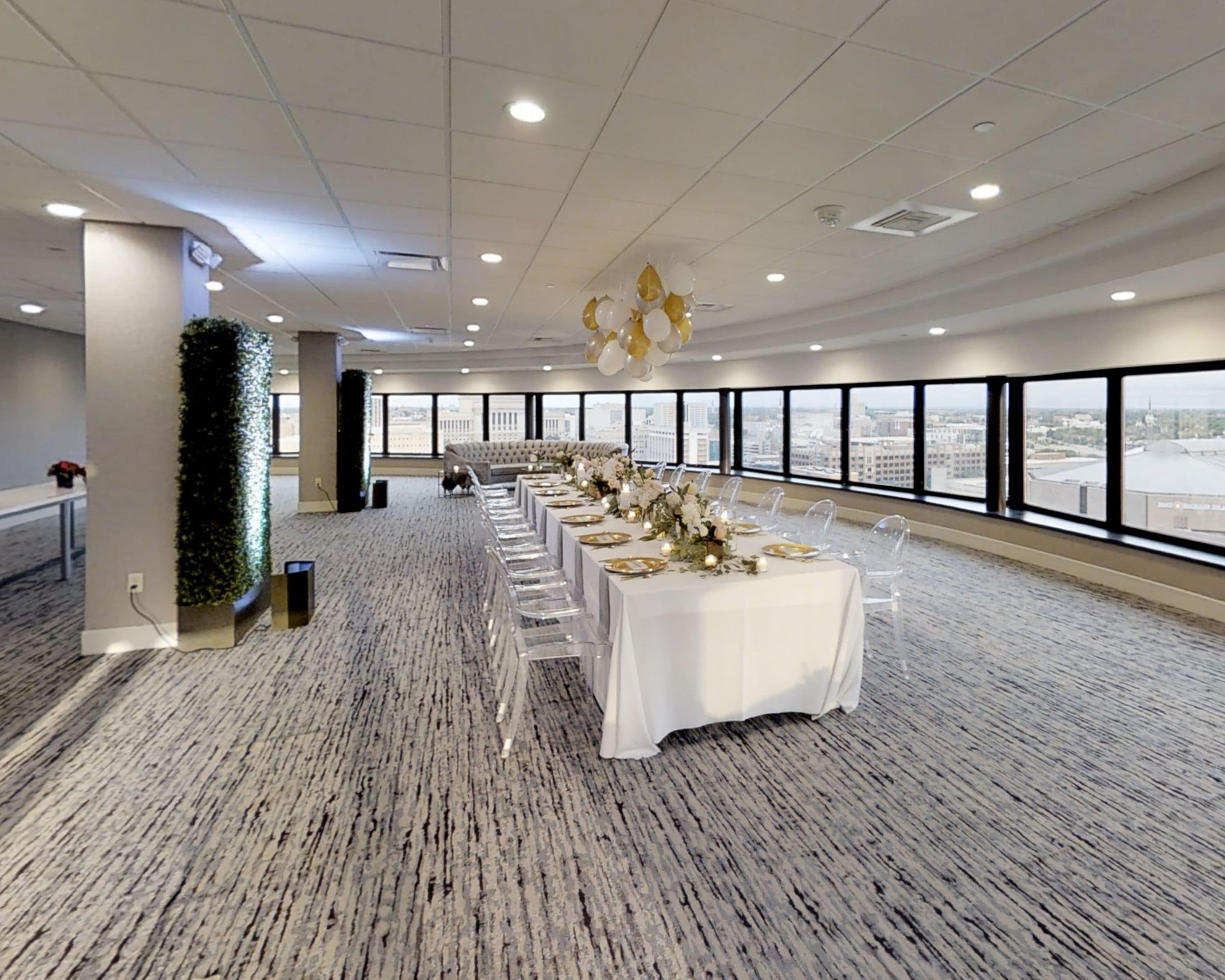 A spacious conference room with a long table set for a formal event, large windows offering a city view, and a decorative ceiling light fixture.