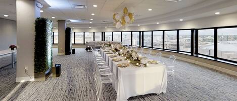 A spacious conference room with a long table set for a formal event, large windows offering a city view, and a decorative ceiling light fixture.