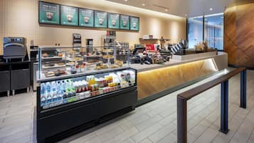 A modern coffee shop with a self-serve counter, refrigerated display case with beverages, and a menu board above.