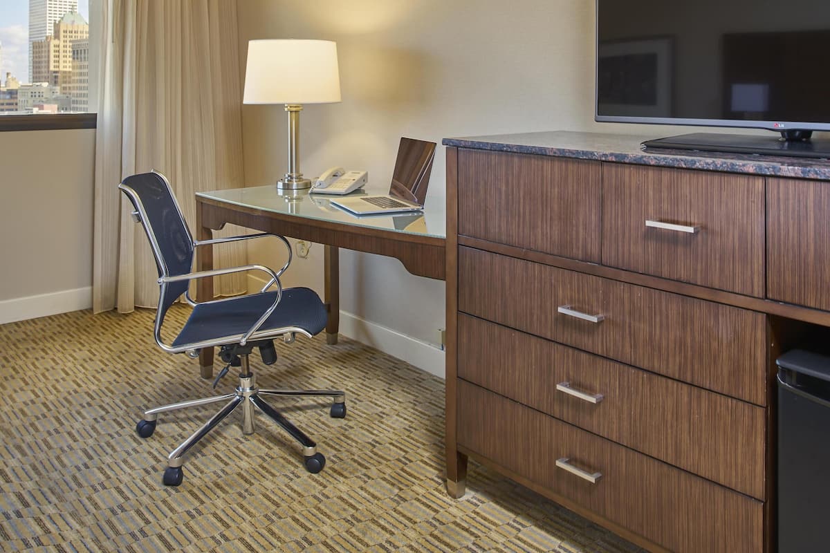 A hotel room with a desk, chair, lamp, TV, and a view of the city.