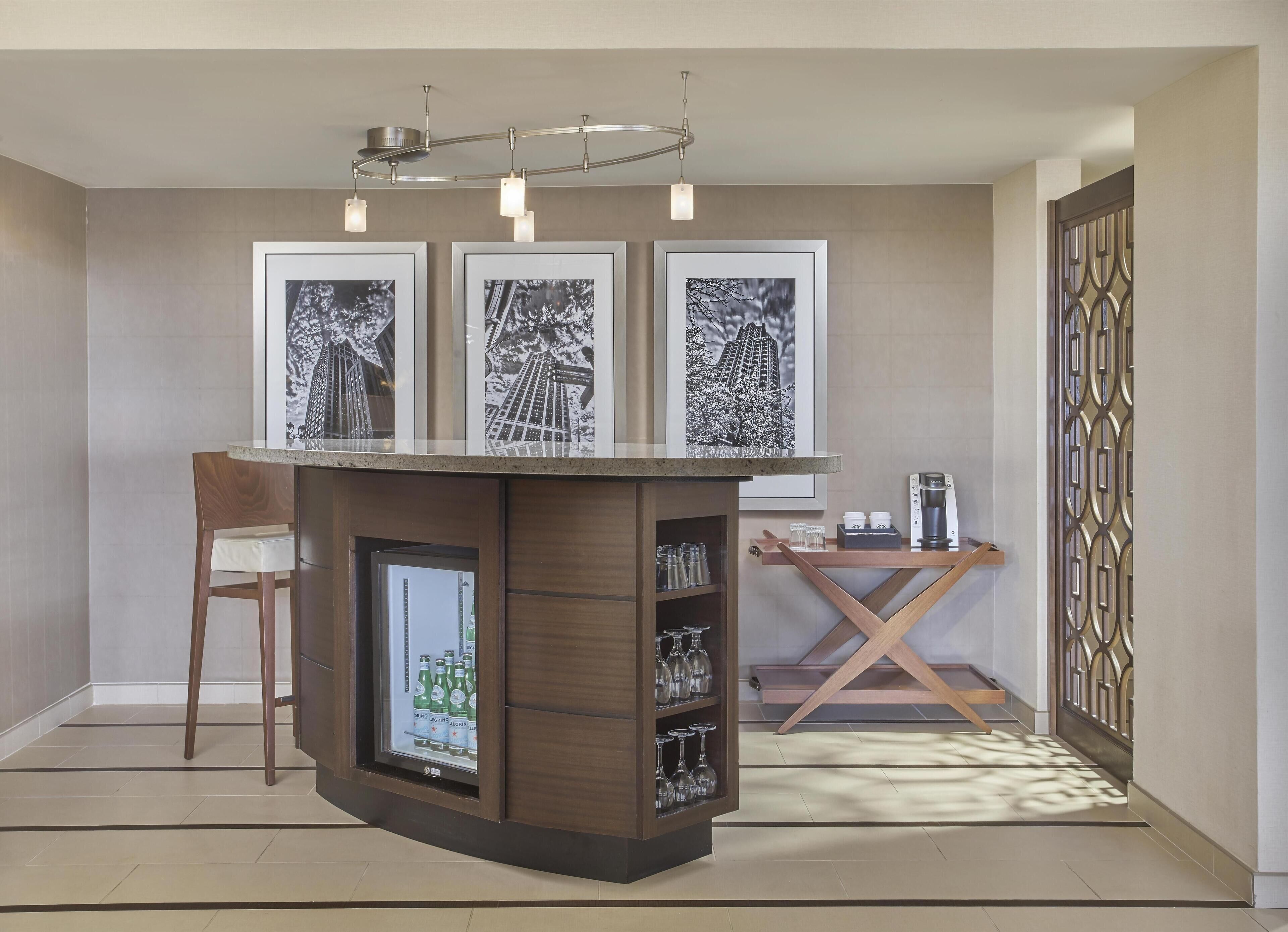 A modern bar area with a wooden counter, a refrigerator with bottles, and framed pictures on the wall.