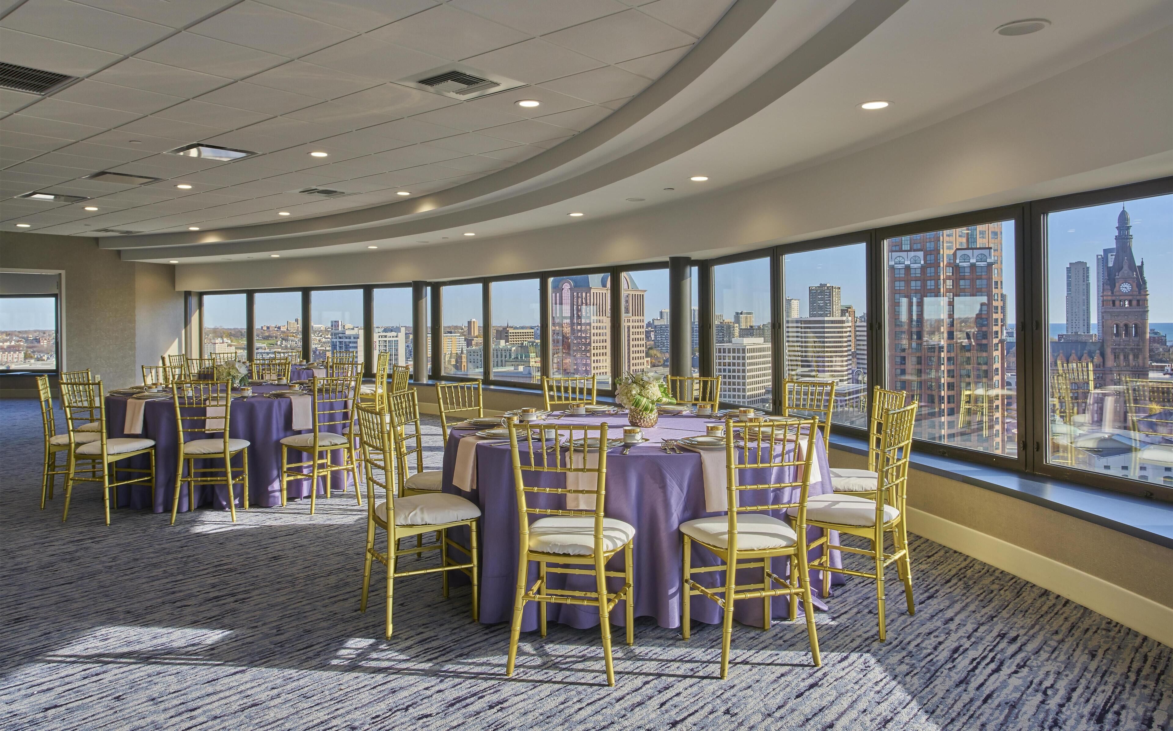 Un salón de conferencias con mesas redondas, sillas doradas y grandes ventanales con vistas a la ciudad.