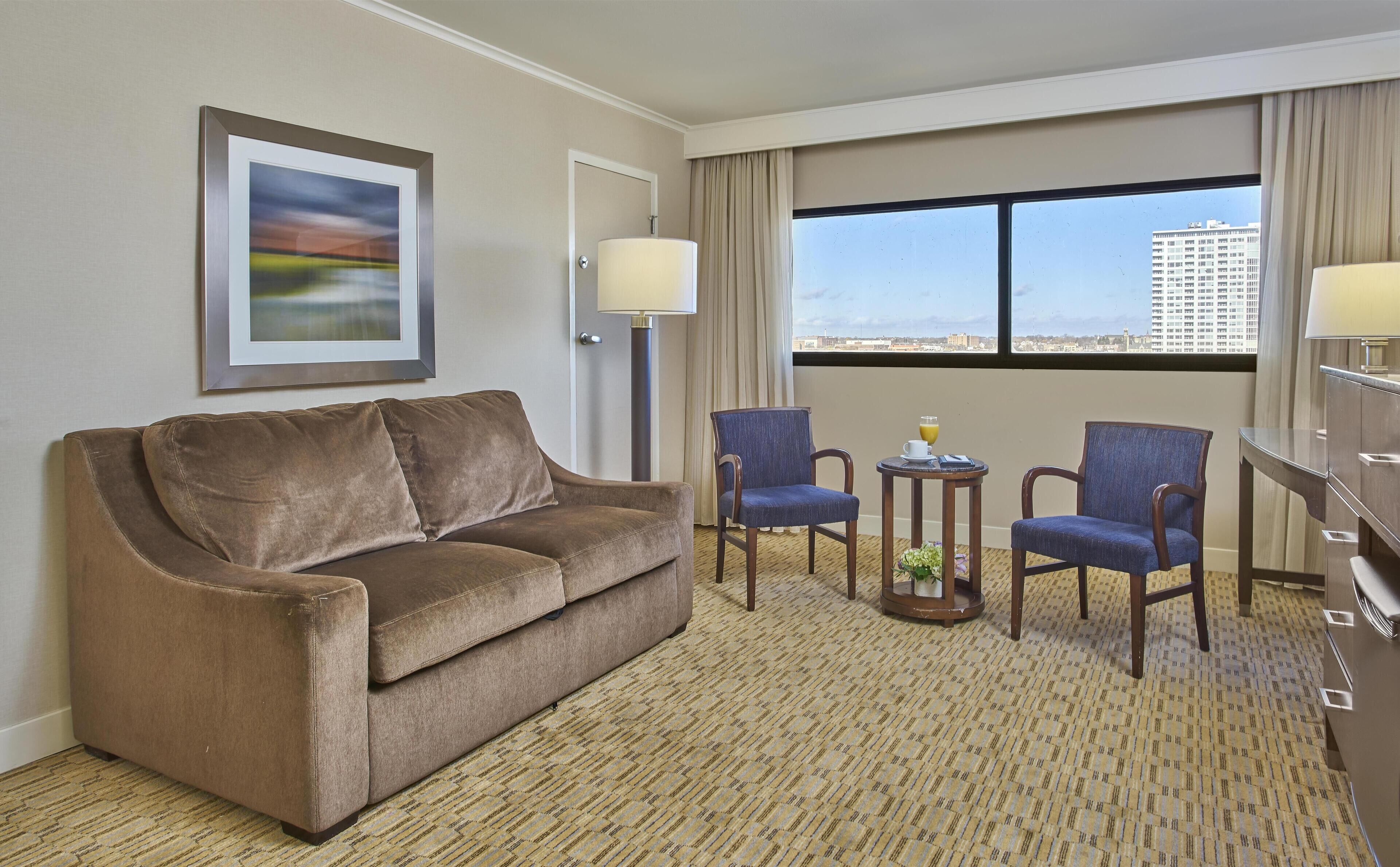 A hotel room with a brown sofa, two blue armchairs, a small table with a lamp, and a large window offering a city view.