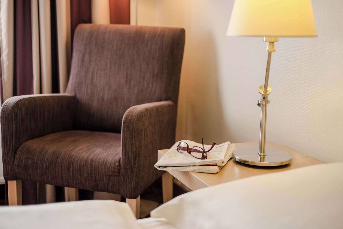 Een hotelkamer met een bruine fauteuil, een nachtkastje met een lamp erop, en een boek met een bril.