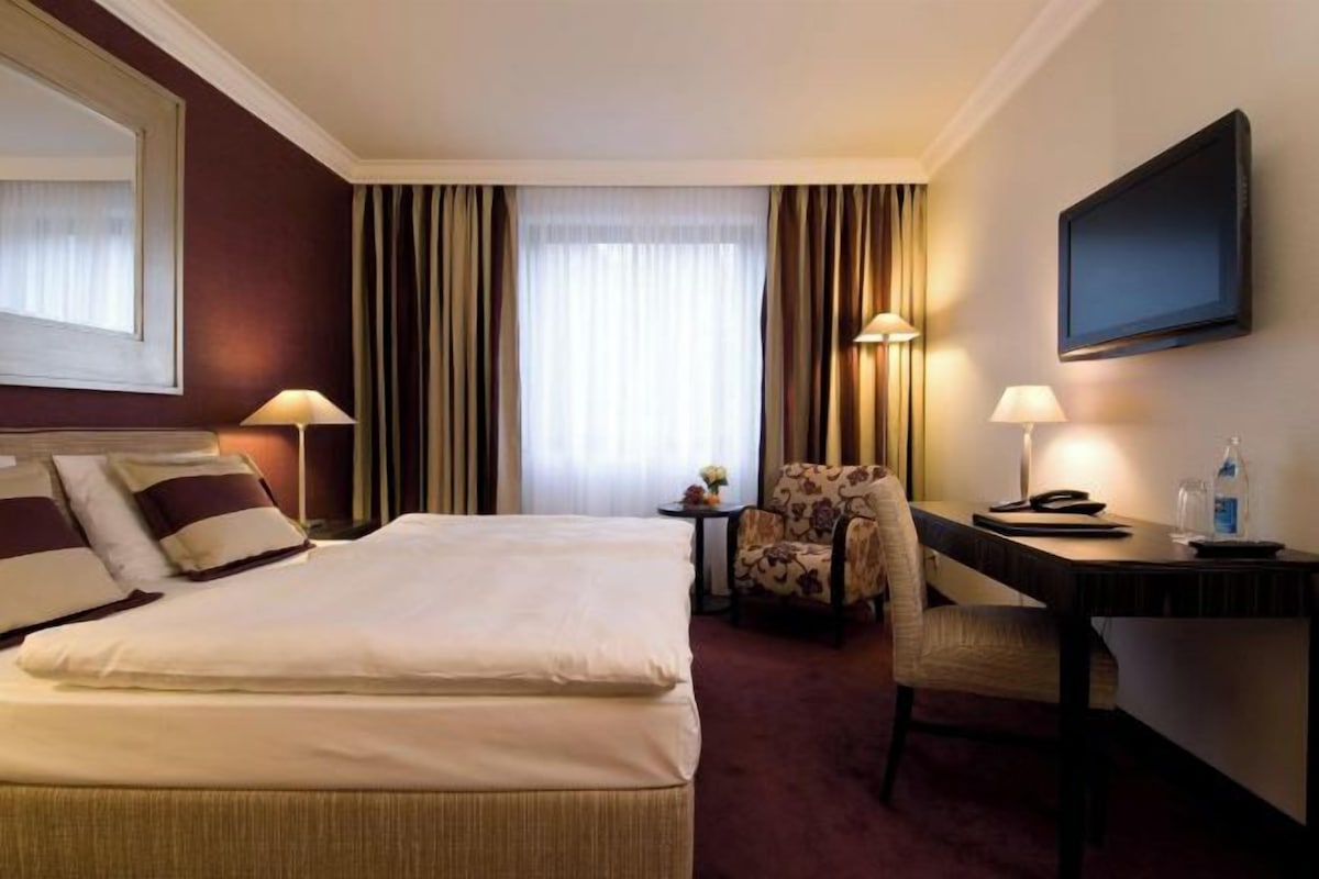 Habitación de hotel con una cama grande, un escritorio con una silla, un televisor y una mesita de noche con una lámpara.