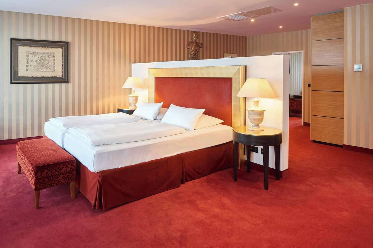 Een hotelkamer met een groot bed, twee nachtkastjes met lampen, een rood vloerkleed, een houten garderobe en een lijst met een afbeelding aan de muur.