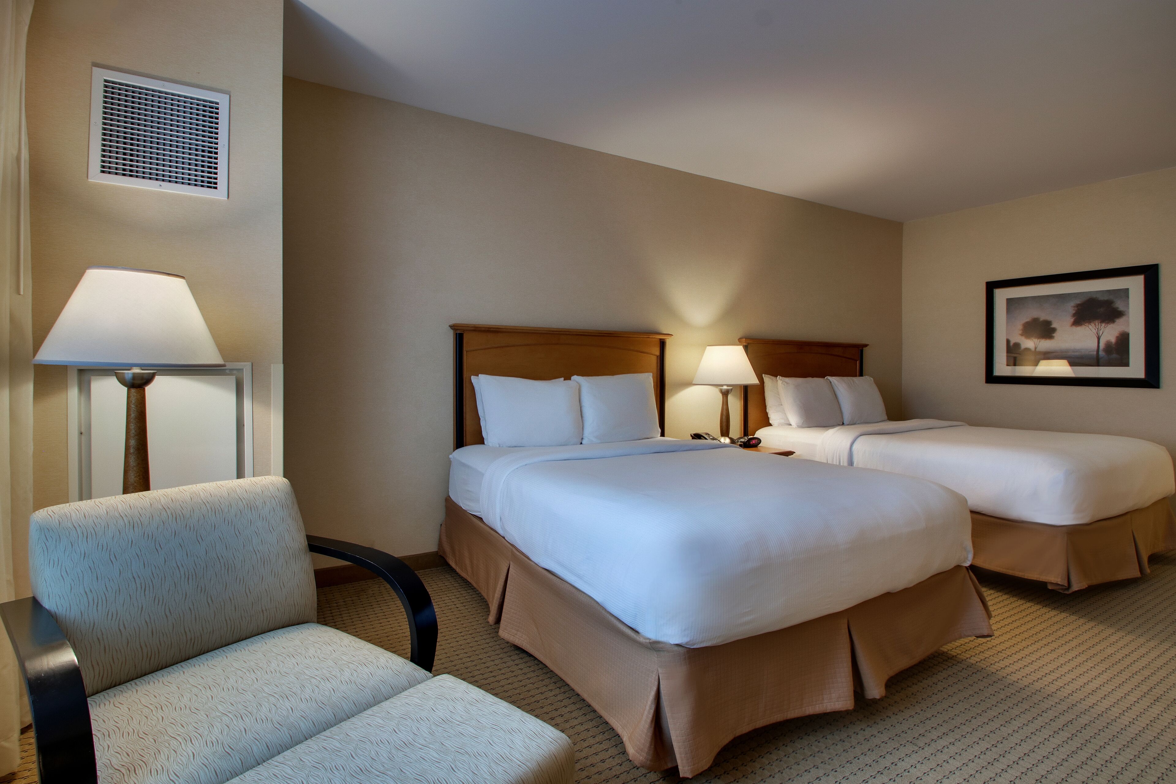 A hotel room with two beds, a chair, a lamp, and a picture on the wall.