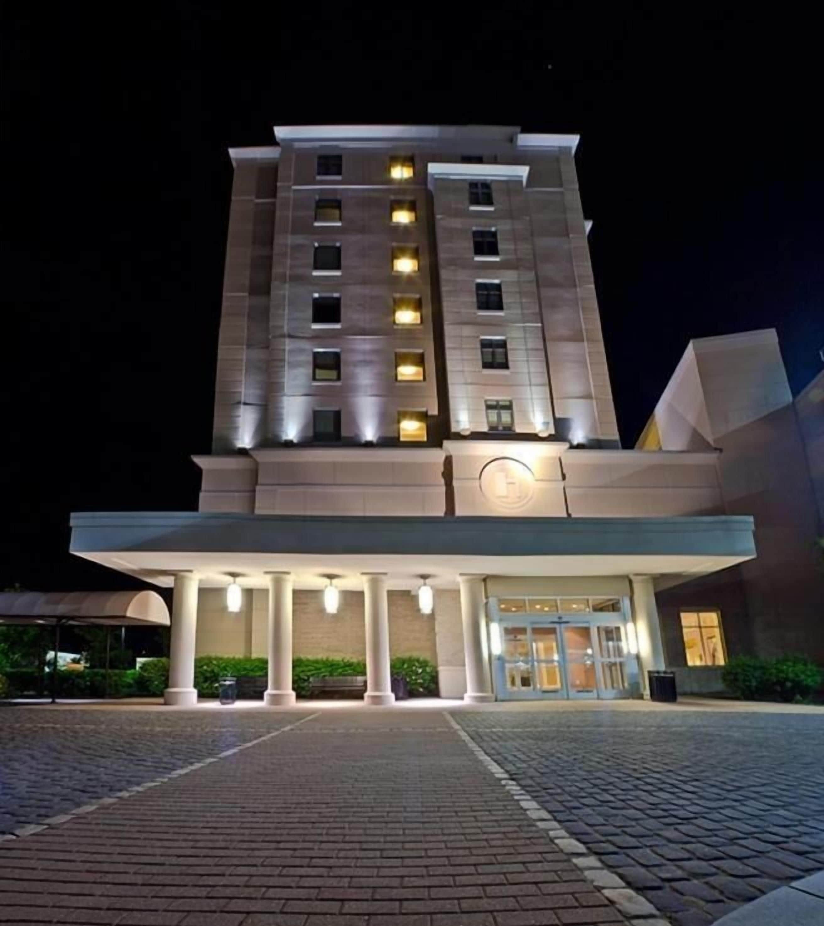 Un hotel de varios pisos con fachada moderna, entrada iluminada y un área cubierta con columnas.