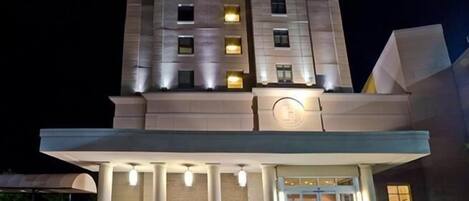 Un hotel de varios pisos con fachada moderna, entrada iluminada y un área cubierta con columnas.